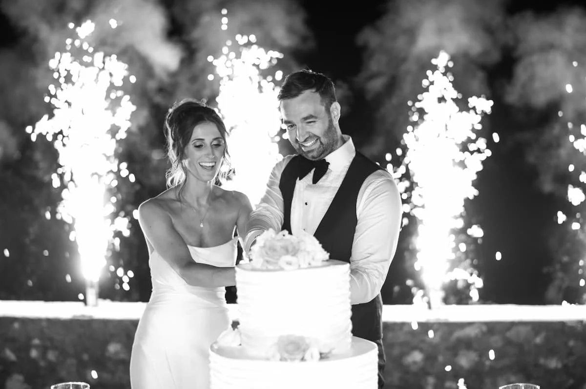 Black and white photograph of a bride and groom smiling while cutting their tiered wedding cake at night, backed by a spectacular display of indoor cold spark fountains (pyrotechnics).