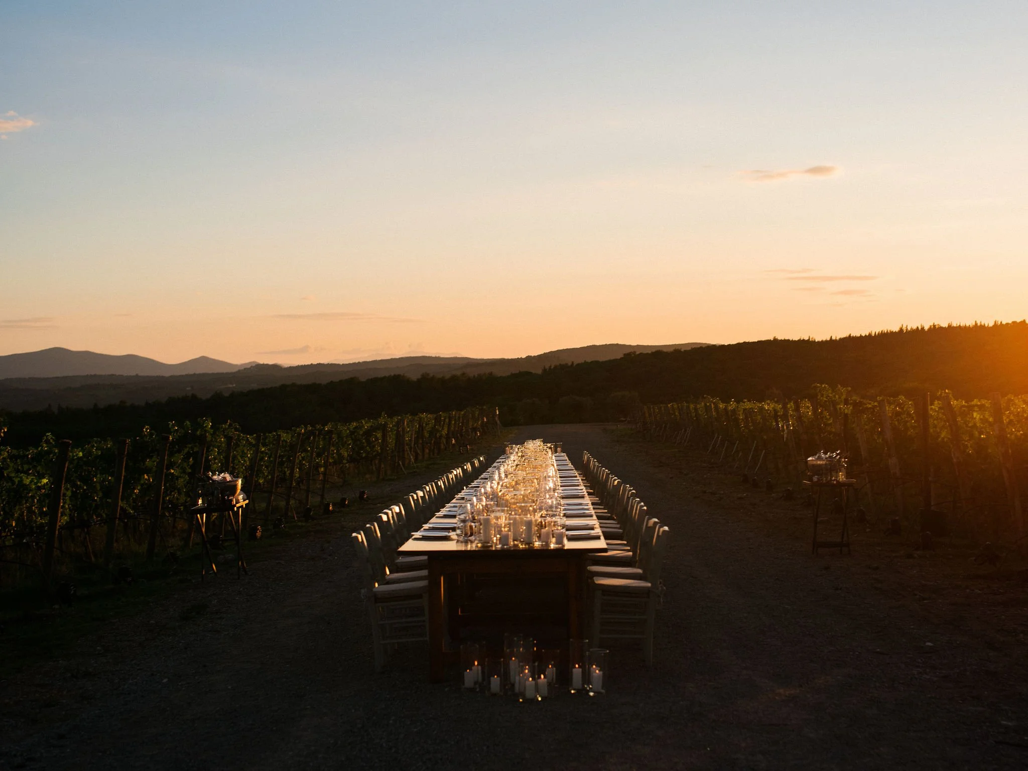 A long, imperial wedding reception table set up between rows of a vineyard in Tuscany during a golden hour sunset, lit by numerous glowing candles.