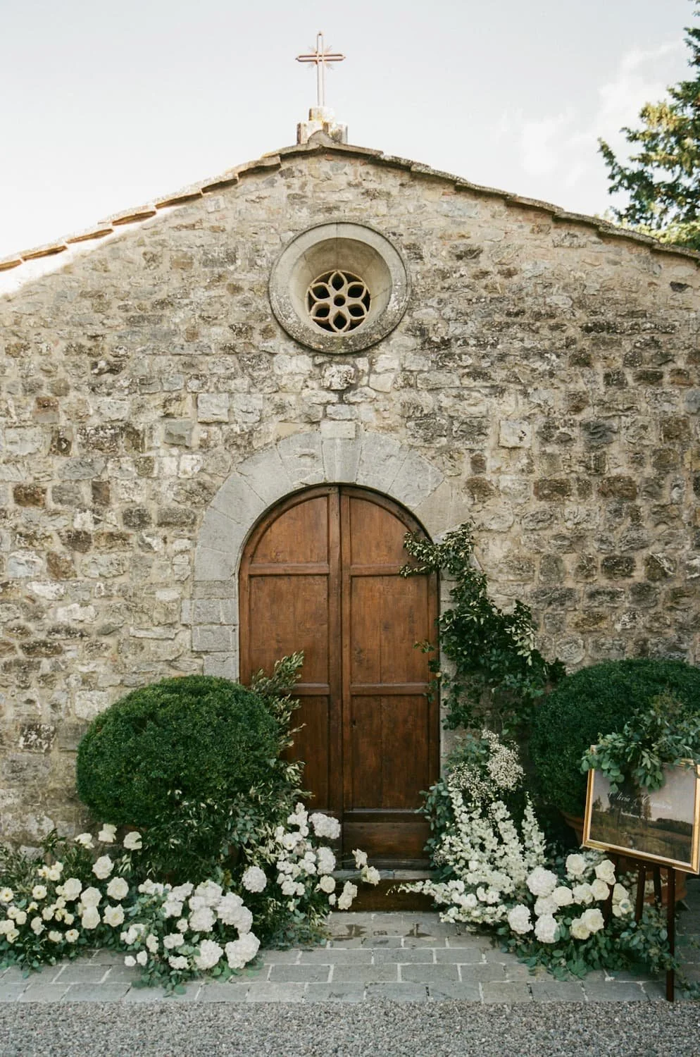 Historic stone chapel facade with arched wooden doors decorated with lush white roses, greenery, and an easel welcome sign at Rosewood Castiglion del Bosco.