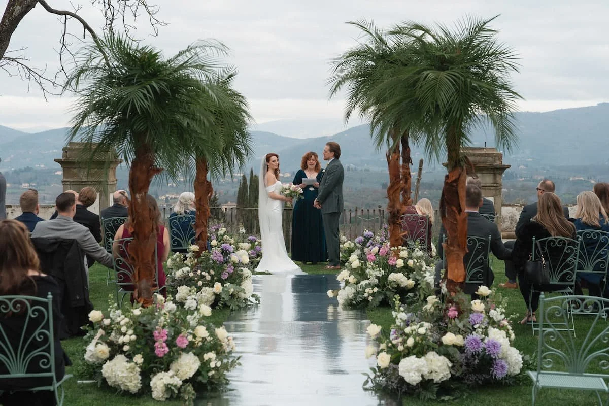 Outdoor winter wedding ceremony in Tuscany featuring a glossy mirrored aisle runner, lush meadow-like floral arrangements with pink, white and green tones, and tall palm trees framing a bride and groom against the misty rolling Italian hills.
