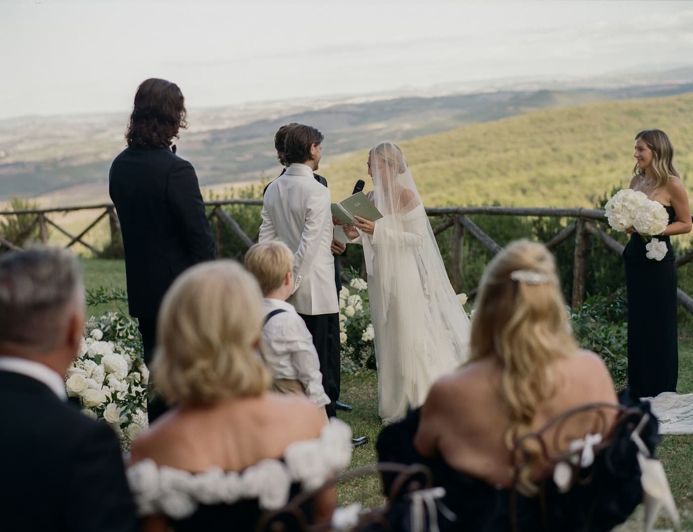 Bride in a flowing veil and off-the-shoulder gown reading vows into a microphone to the groom in a cream tuxedo jacket overlooking the Tuscan hills at Rosewood Castiglion del Bosco.