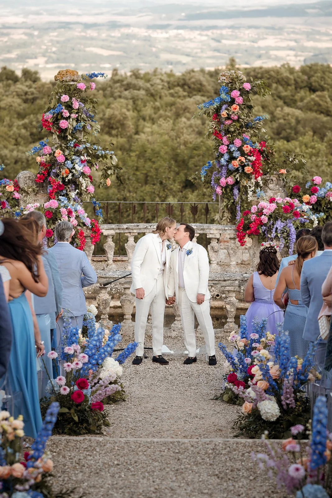 Same-sex couple kissing during a luxury destination wedding ceremony decorated with vibrant floral arches at a historic Tuscan estate in Italy.