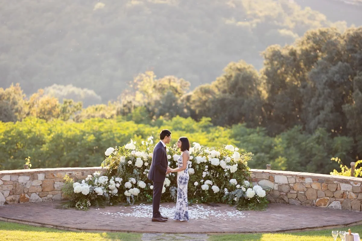 A romantic couple standing together on a private stone terrace in Tuscany, framed by lush white floral installations and rolling green hills at sunset