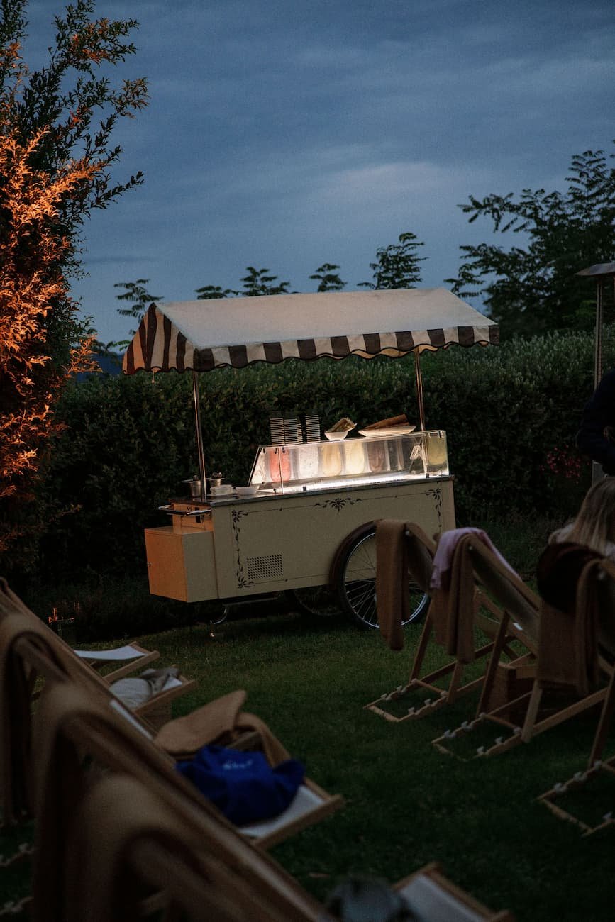 Traditional vintage Italian gelato cart with a striped canopy lit at dusk on a lawn during a wedding evening event at Rosewood Castiglion del Bosco.