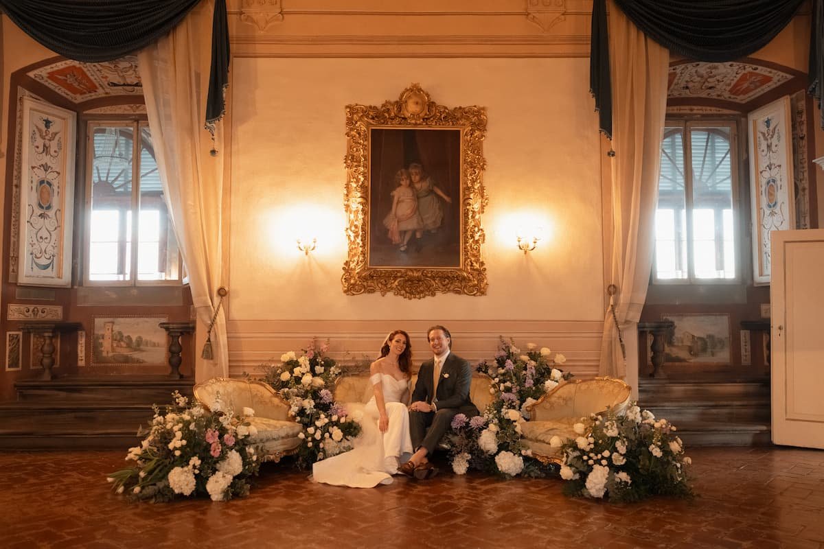 A bride and groom sitting on an antique gold velvet sofa in a luxury Tuscan villa hall, framed by lush, grounded lavender and white floral arrangements beneath a massive gilded portrait painting.