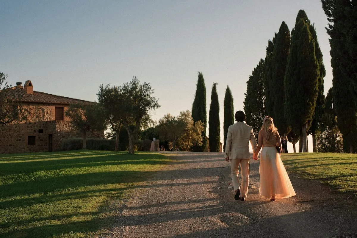 Bride and groom holding hands while walking down a gravel pathway lined with cypress trees and a rustic stone farmhouse during golden hour at Rosewood Castiglion del Bosco.