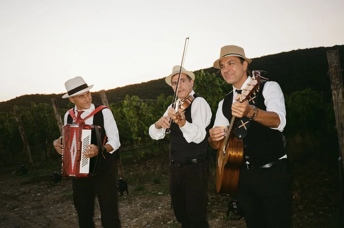Three traditional Italian folk musicians in fedora hats, waistcoats, and ties playing accordion, violin, and acoustic guitar outdoors by the vineyard at dusk.