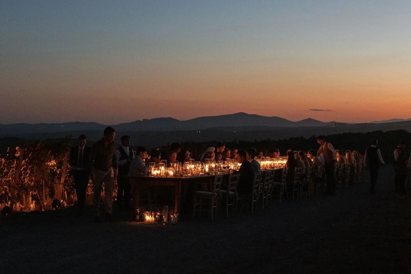Long wooden banquet table lit exclusively with glowing pillar candles for a vineyard welcome dinner at dusk overlooking the Tuscan hills at Rosewood Castiglion del Bosco.