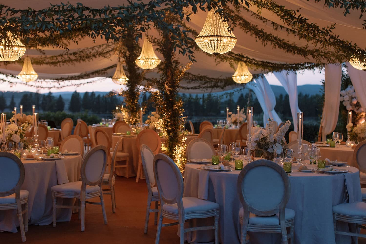 Elegantly styled outdoor wedding reception marquee at dusk featuring glowing crystal chandeliers, greenery-wrapped poles, round dining tables, and Louis XVI-style chairs at Rosewood Castiglion del Bosco.
