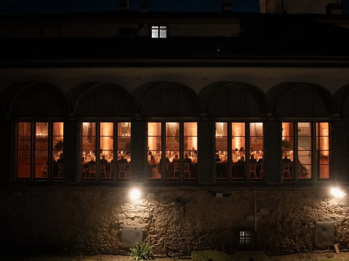 Exterior night view of an elegant candlelight wedding reception dinner held in an illuminated historic Tuscan palazzo.