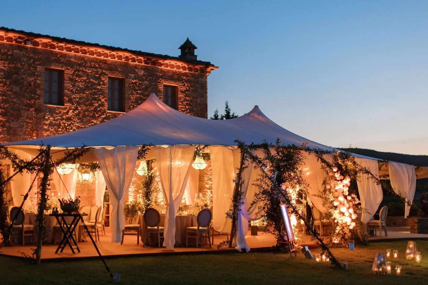 Exterior view at twilight of a grand white sailcloth marquee tent with draped fabric, glowing chandeliers, and floral arches set against an illuminated stone villa at Rosewood Castiglion del Bosco.
