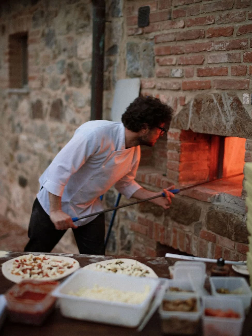 A chef sliding fresh pizza into a glowing brick wood-fired oven outdoors alongside pizza prep ingredients at Rosewood Castiglion del Bosco.