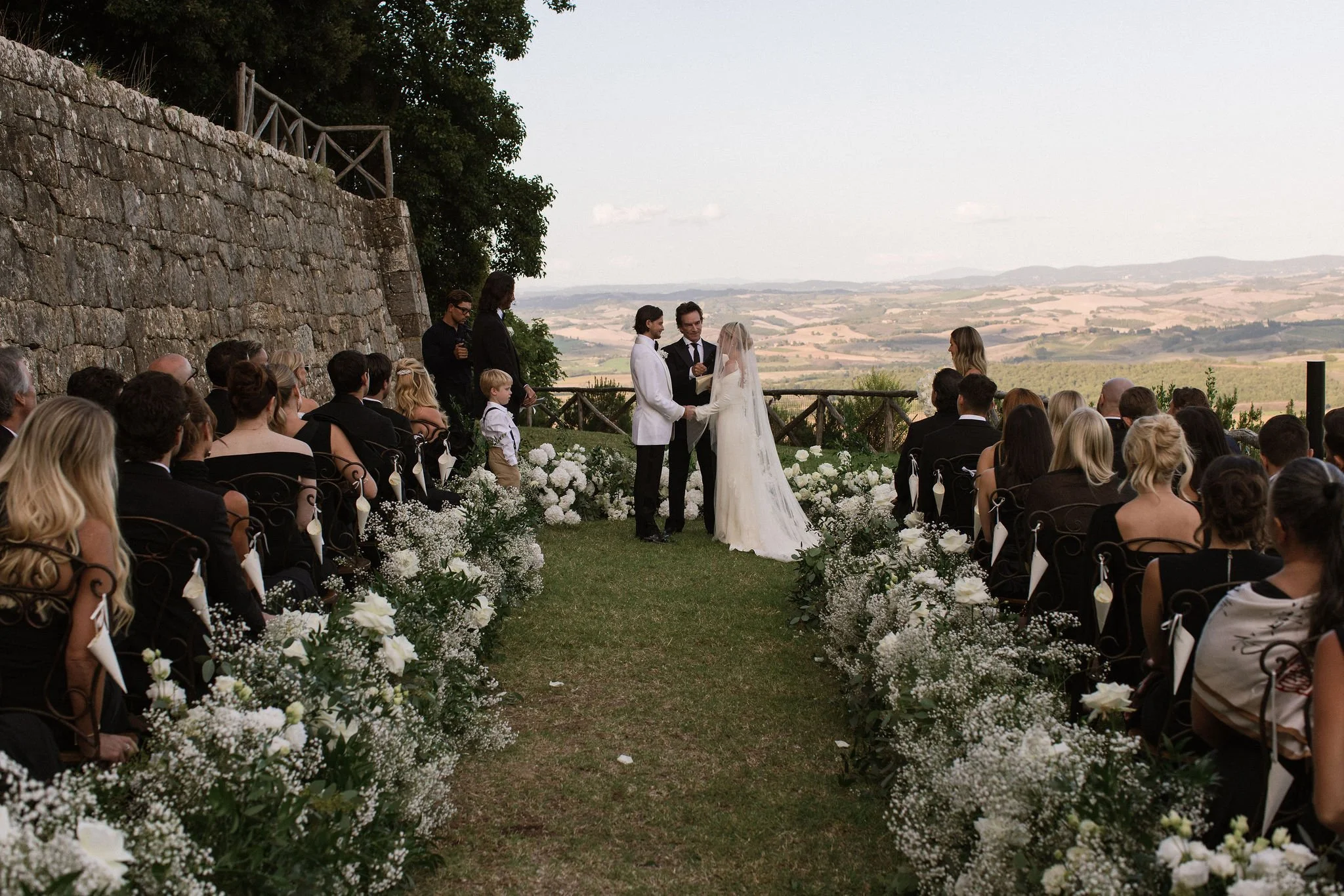 Wide shot of a luxury destination wedding ceremony in Tuscany, featuring a bride and groom at the altar on a grassy lawn bordered by lush white flowers, overlooking panoramic rolling hills and an ancient stone wall.
