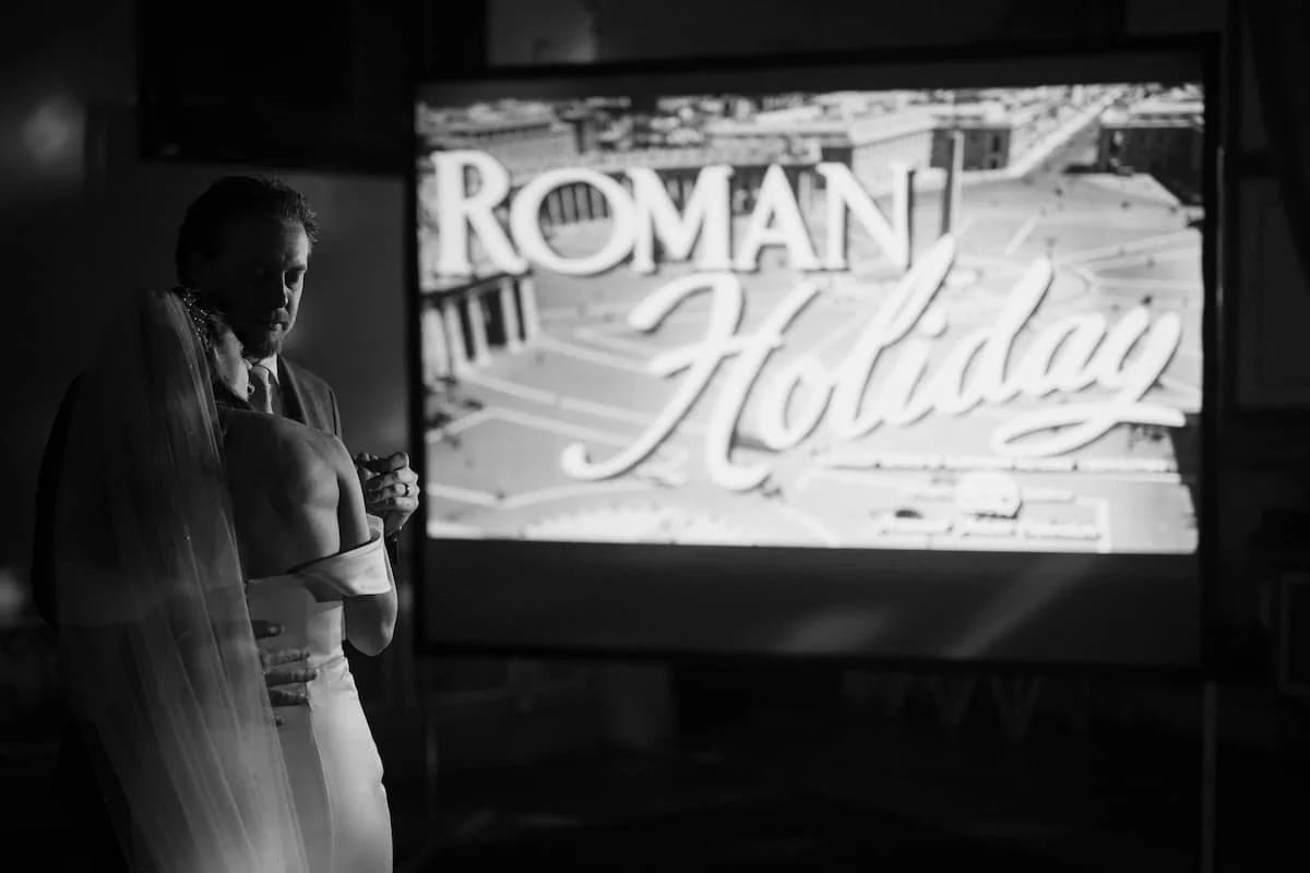 Bride and groom slow dancing at an indoor wedding reception in Tuscany with classic Roman Holiday movie playing on a projector screen behind them.