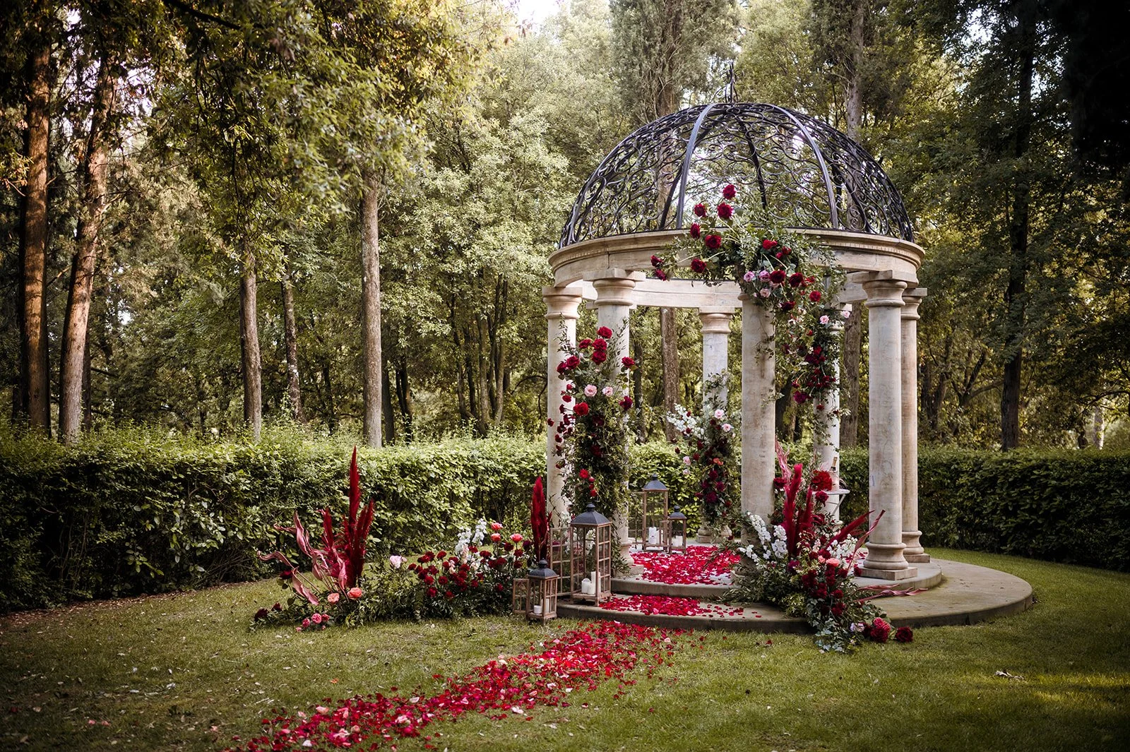 An ornate stone wedding gazebo with a black wrought iron dome roof in a lush Tuscan forest garden, decorated with deep red roses, pampas grass, lanterns, and a path of red flower petals.