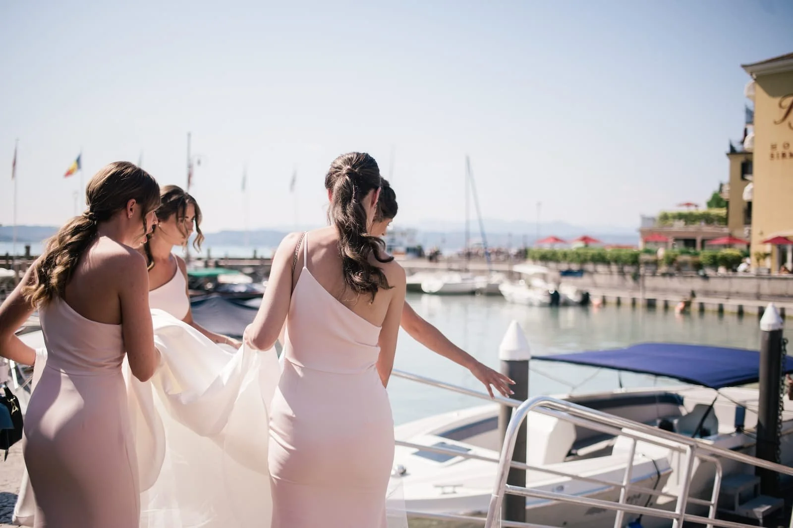 Bridesmaids in elegant blush pink dresses holding up a bridal gown train while boarding a luxury boat at a scenic lakeside harbor town for a destination wedding.