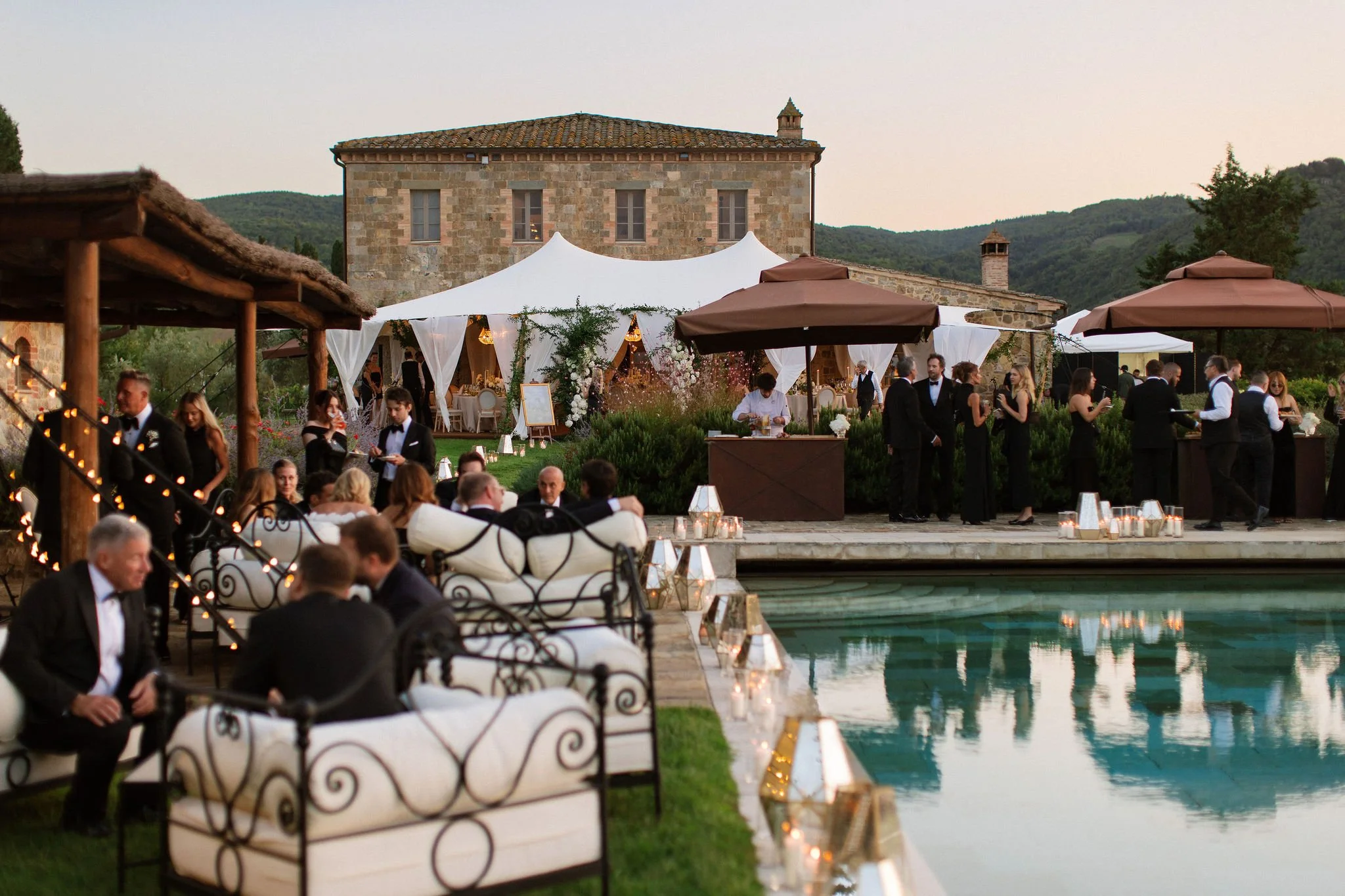 Luxury outdoor wedding reception at sunset featuring a poolside cocktail hour, elegant guests, catering staff, a sailcloth tent, and lighting design at a historic stone villa estate.