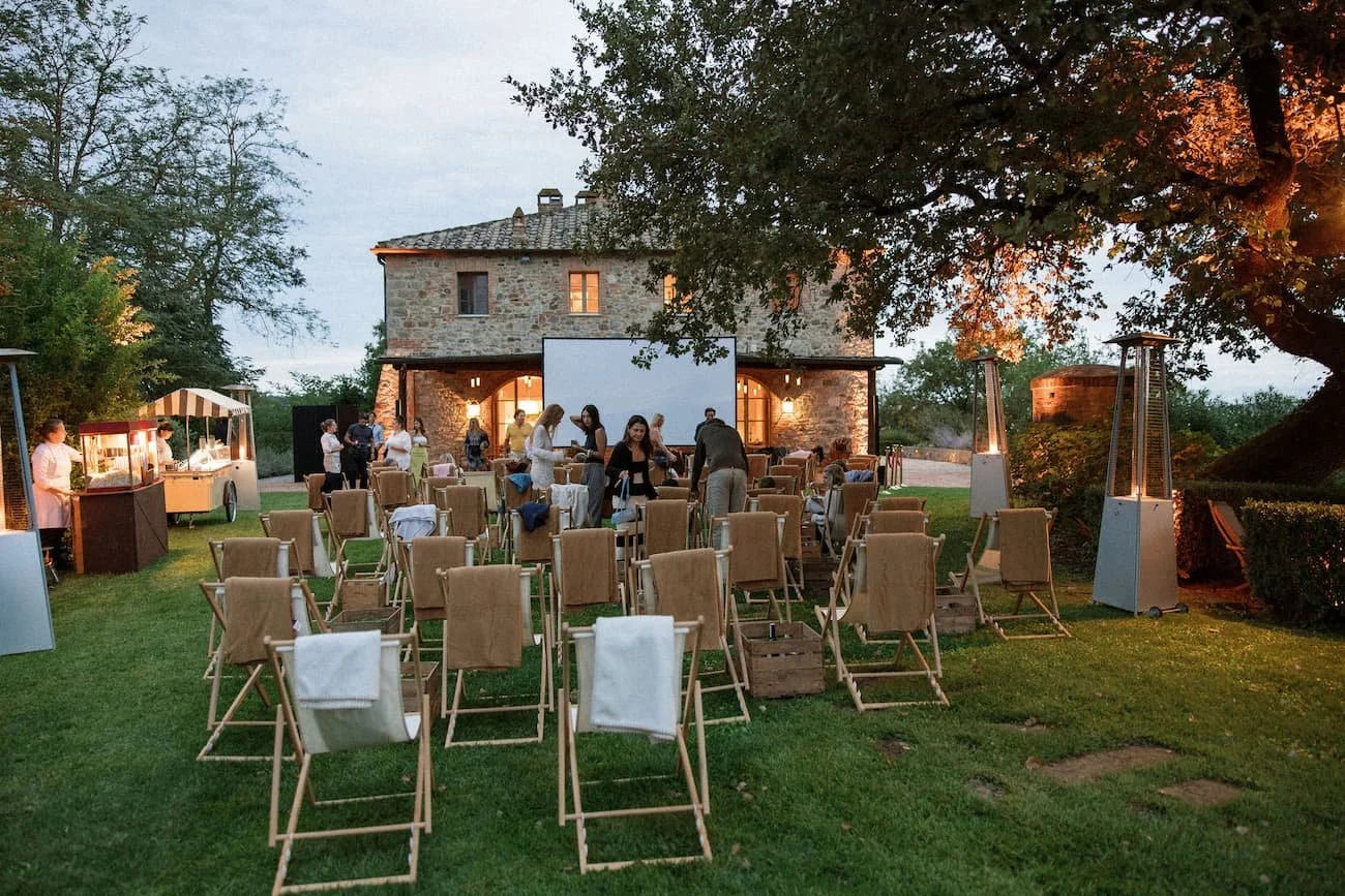 Wide shot of an outdoor movie night setup on the lawn in front of a stone villa, featuring a large projector screen, deck chairs, patio heaters, popcorn, and gelato carts at dusk at Rosewood Castiglion del Bosco.