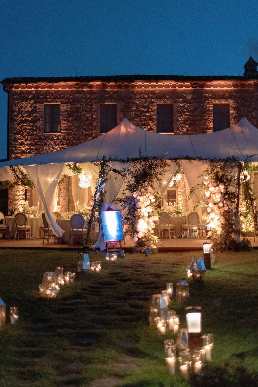 Lawn pathway lined with glowing glass lanterns leading to a warmly lit reception marquee tent beside a stone villa at night at Rosewood Castiglion del Bosco.
