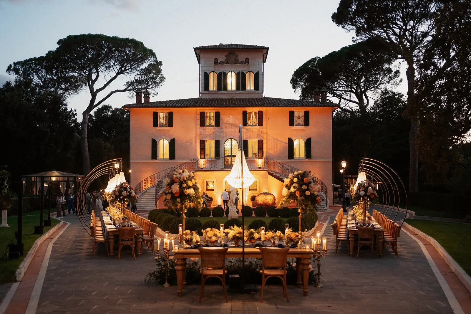 An outdoor luxury wedding reception at twilight in front of an illuminated historic Italian villa in Tuscany, featuring arched lighting installations over long dinner tables, floral centerpieces, and a glowing crystal chandelier.