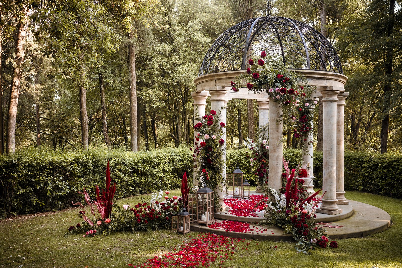 A luxurious outdoor marriage proposal setup inside a classical stone gazebo in a lush Tuscan garden, beautifully decorated with deep red roses, romantic lanterns, and a path of red rose petals.