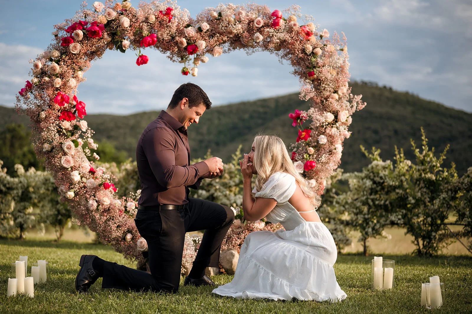 A romantic outdoor marriage proposal in Tuscany featuring a man on one knee proposing under a stunning heart-shaped floral arch decorated with pink roses.