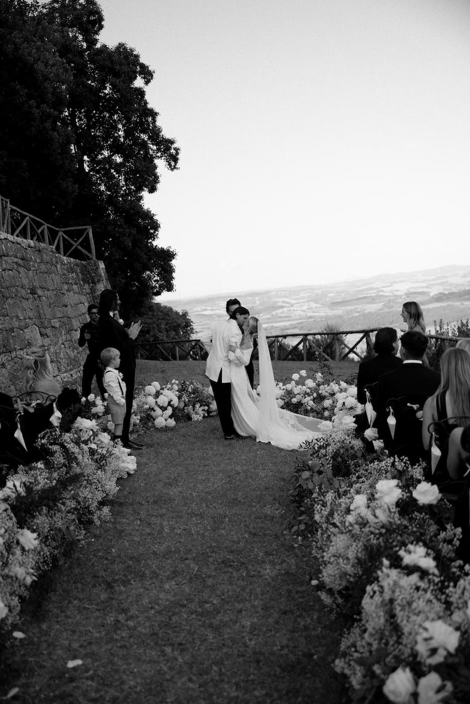 Black and white photograph of the bride and groom sharing their first kiss at the end of the aisle framed by white flowers overlooking the Tuscan hills at Rosewood Castiglion del Bosco.