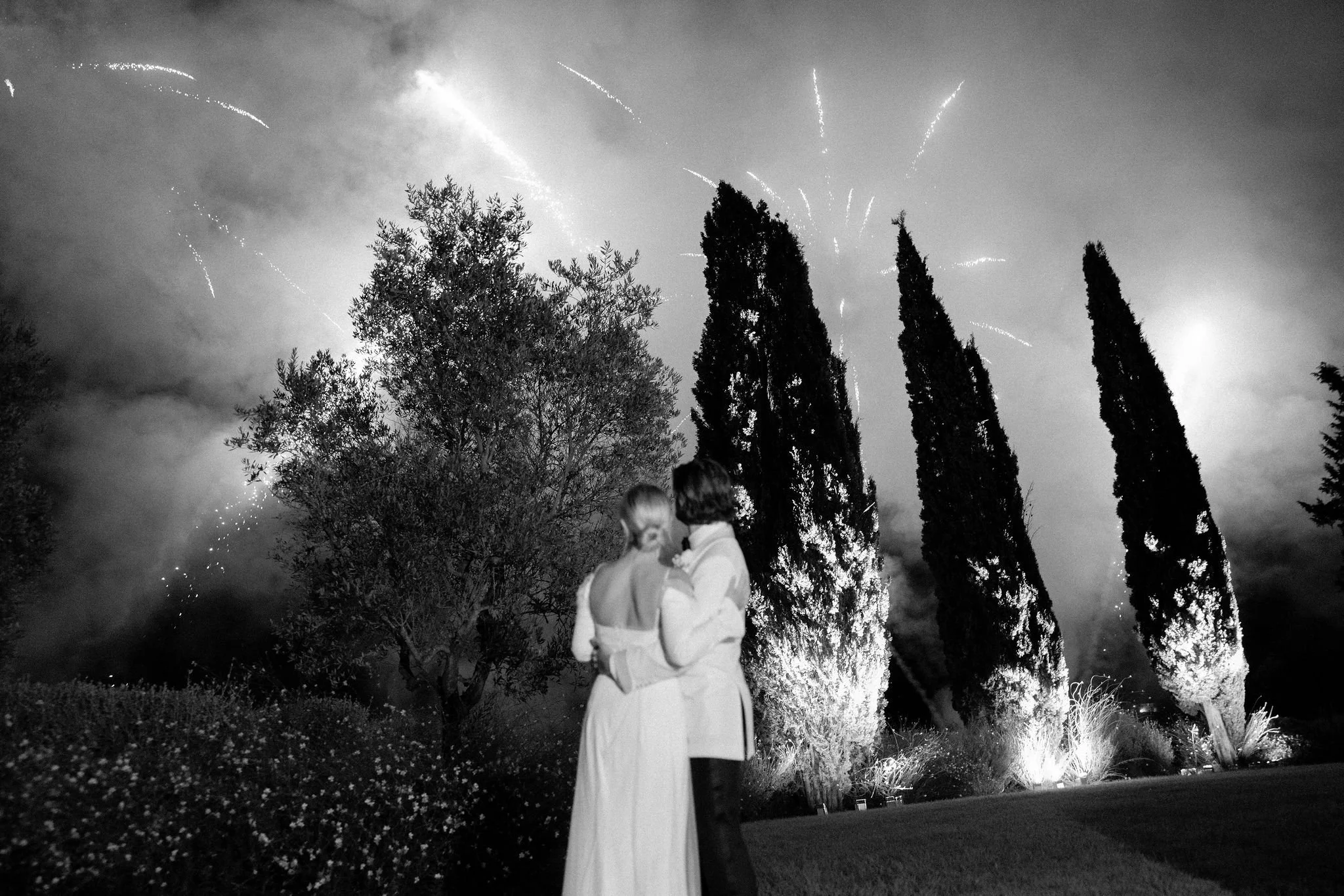 Black and white photograph of a bride and groom embracing from behind, watching a spectacular wedding fireworks display light up the night sky behind tall cypress trees at a luxury estate.