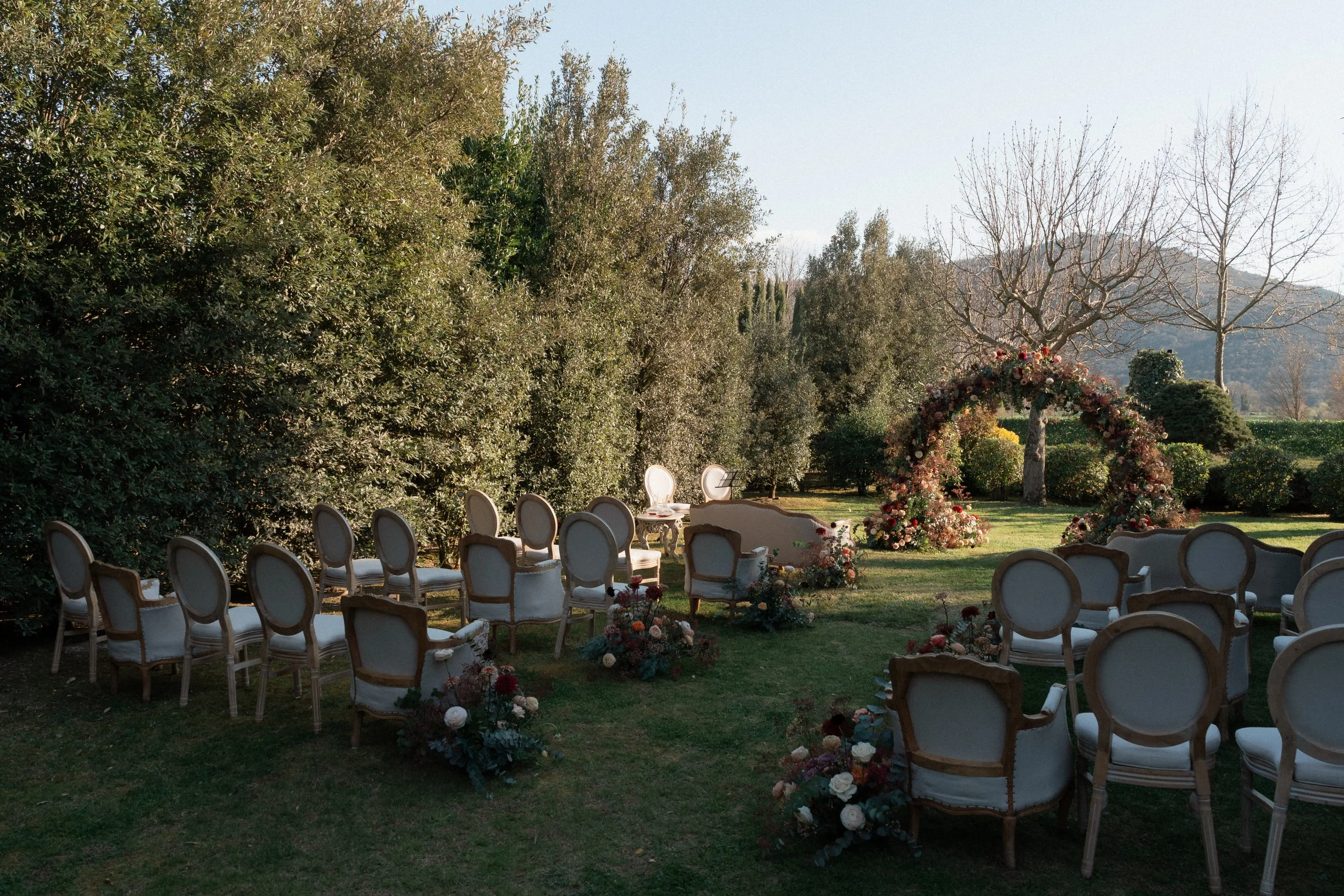 An intimate luxury outdoor wedding ceremony set up on a secluded green lawn in Tuscany, featuring vintage Louis XVI-style upholstered armchairs for guests, moody autumnal floral arrangements, and a circular flower arch nestled against mature trees.