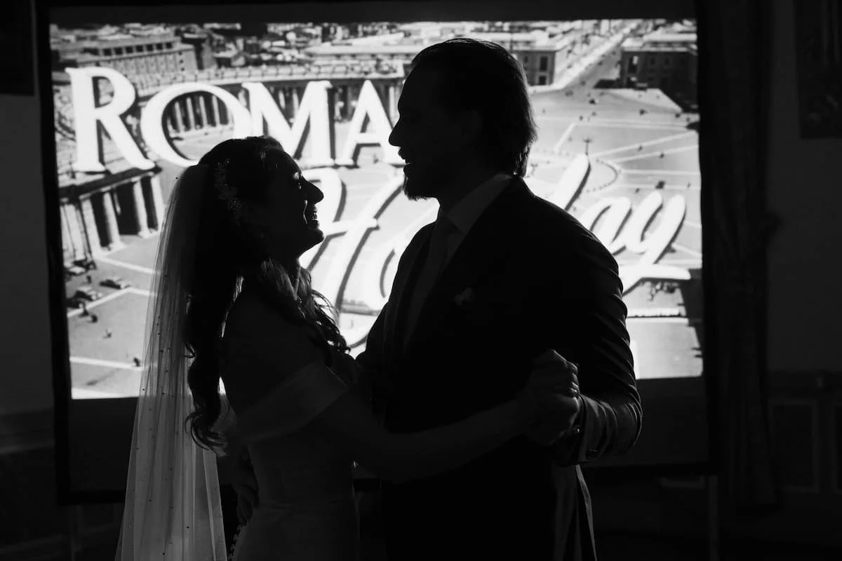 Silhouette of a bride and groom sharing a first dance in front of a vintage "Roma Holiday" film projection during a cozy, indoor winter wedding reception in Tuscany.