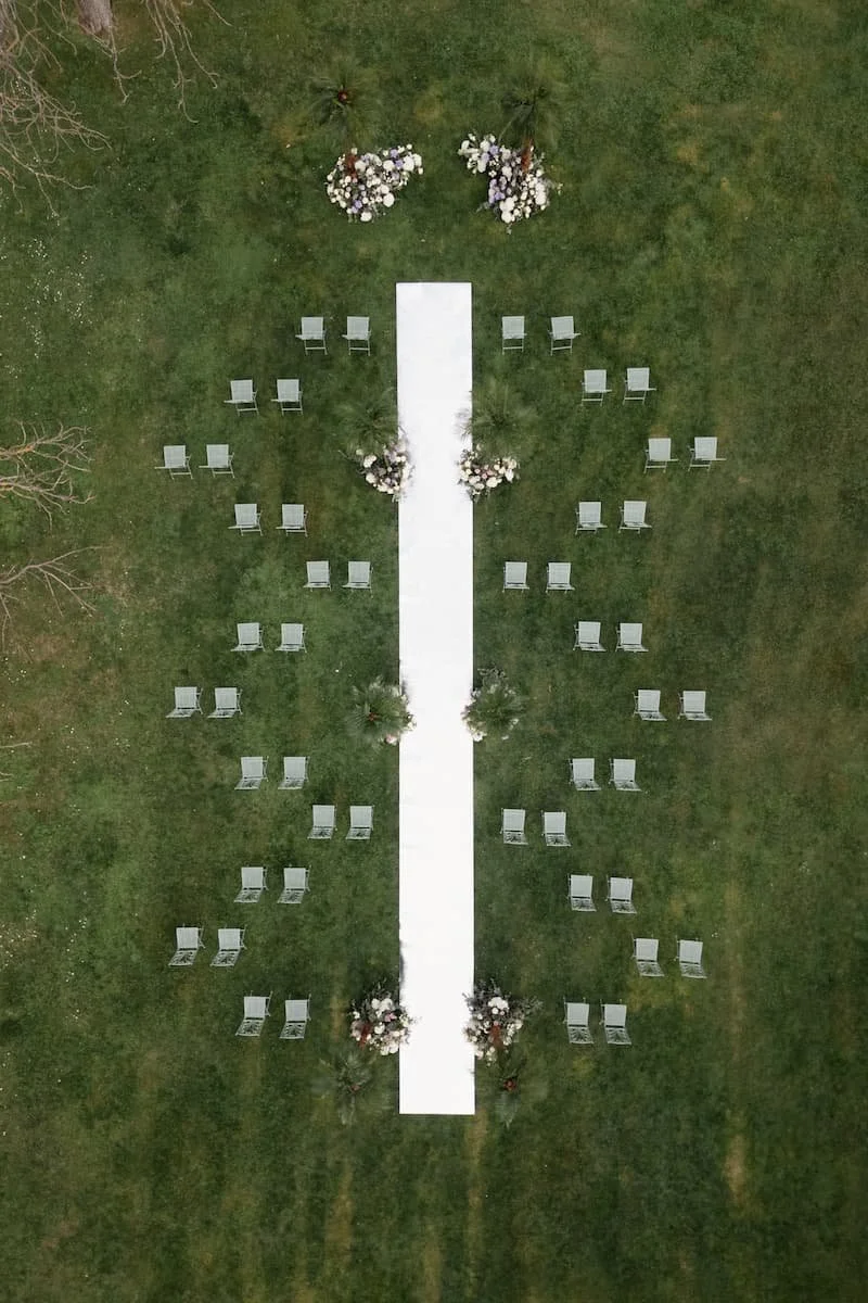 Aerial top-down view of an outdoor wedding ceremony lawn layout with a mirrored aisle, guest chairs, and floral clusters.