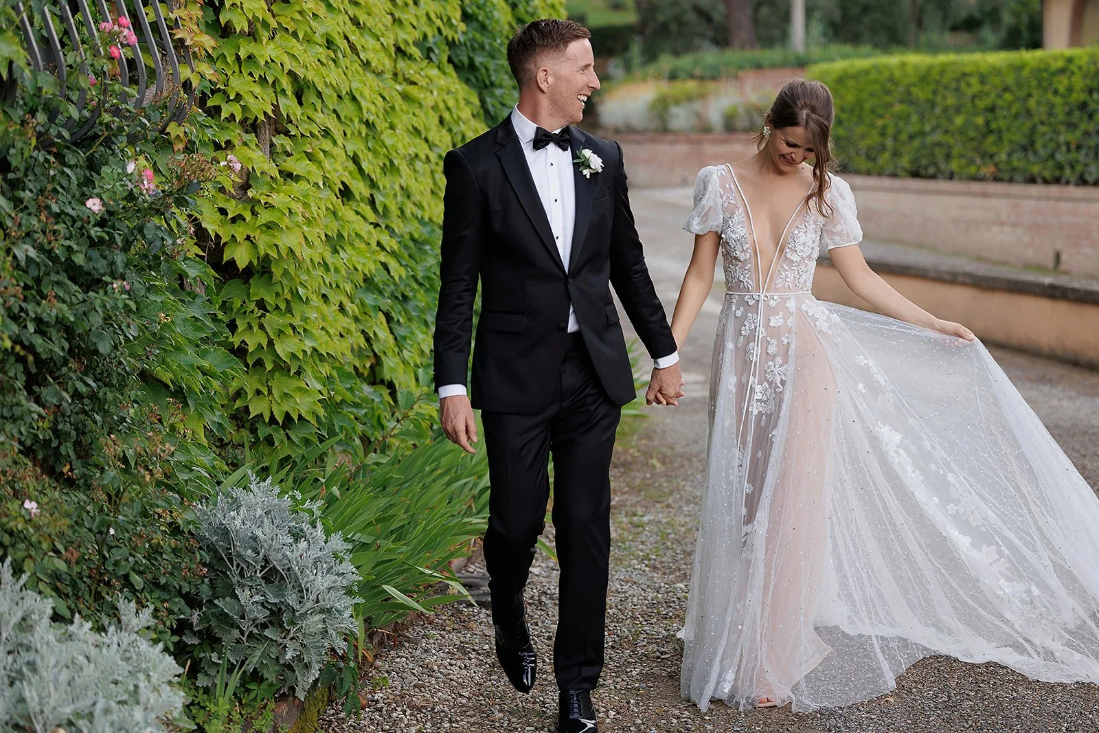 A smiling bride in a sheer, intricately embroidered floral tulle wedding gown holds hands and walks down a gravel pathway with her groom in a classic black tuxedo next to an ivy-covered stone wall.