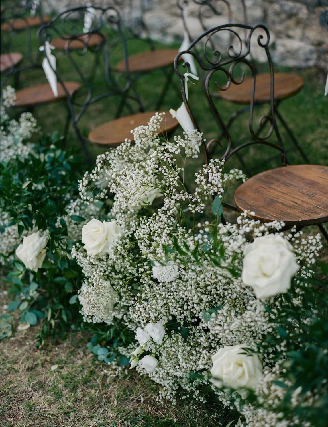 Ground-level wedding ceremony aisle flowers featuring white roses, baby's breath, and greenery bordering wrought-iron chairs with wooden seats on a grass lawn.