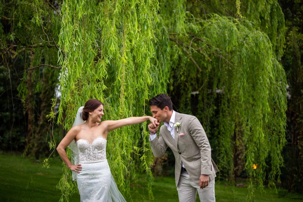A groom in a light beige linen suit kisses the hand of his smiling bride in a strapless lace gown during their outdoor wedding portrait session under a weeping willow tree.