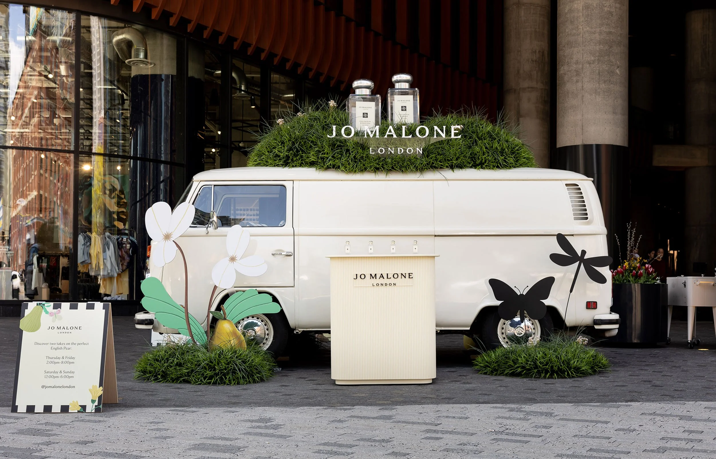McWood custom Jo Malone pop-up featuring a vintage VW van with oversized grassy mounds, giant perfume bottles, flowers, and insects at The Well in Toronto.