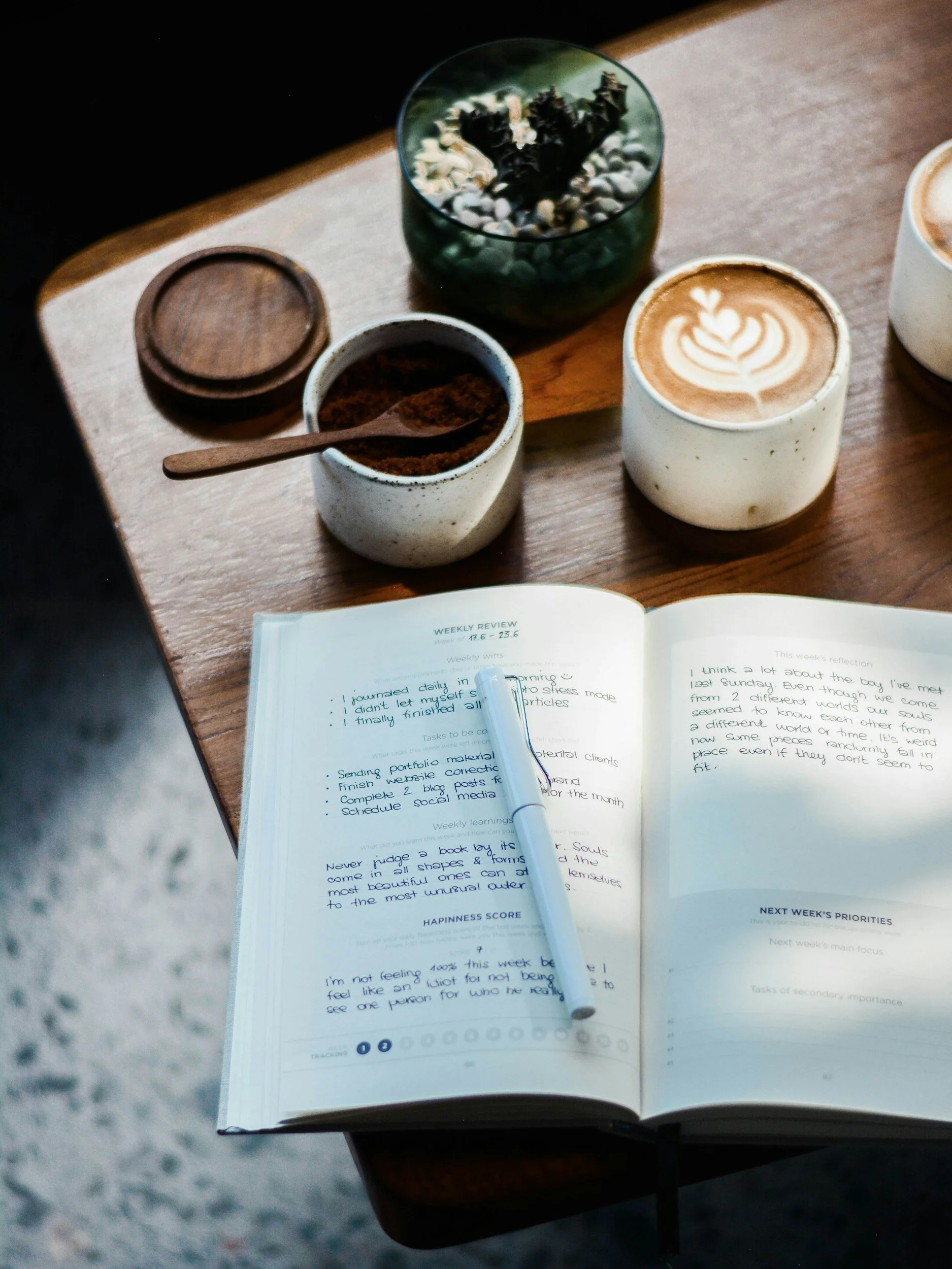 Open planner or notebook with handwritten notes, a blue pen, a cup of latte with latte art, a small container of ground coffee, and a glass pot with decorative rocks and plant inside on a wooden table.