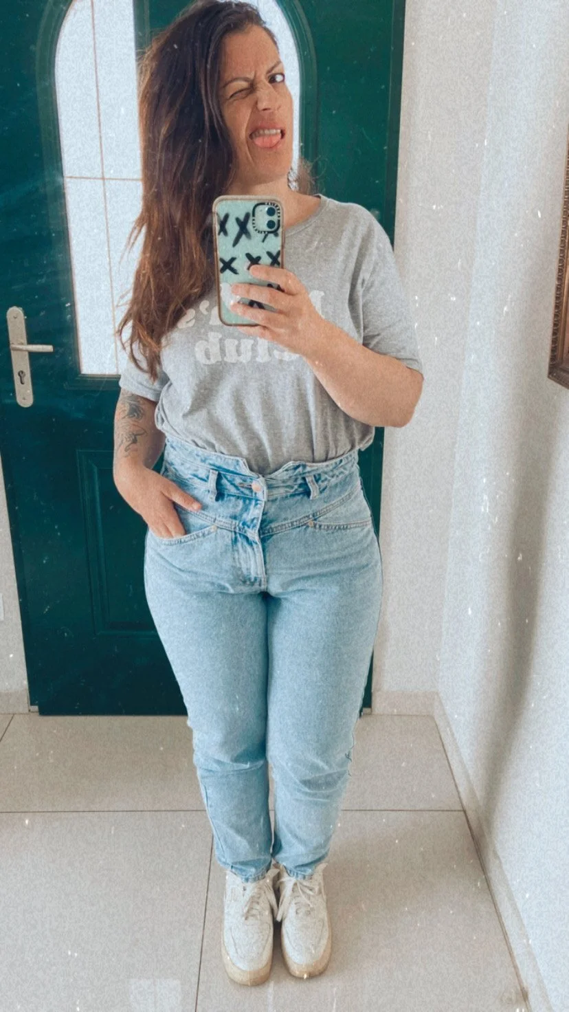 A woman taking a mirror selfie, winking and scrunching her nose, wearing a gray T-shirt, light blue jeans, and white sneakers, standing in front of a green door.