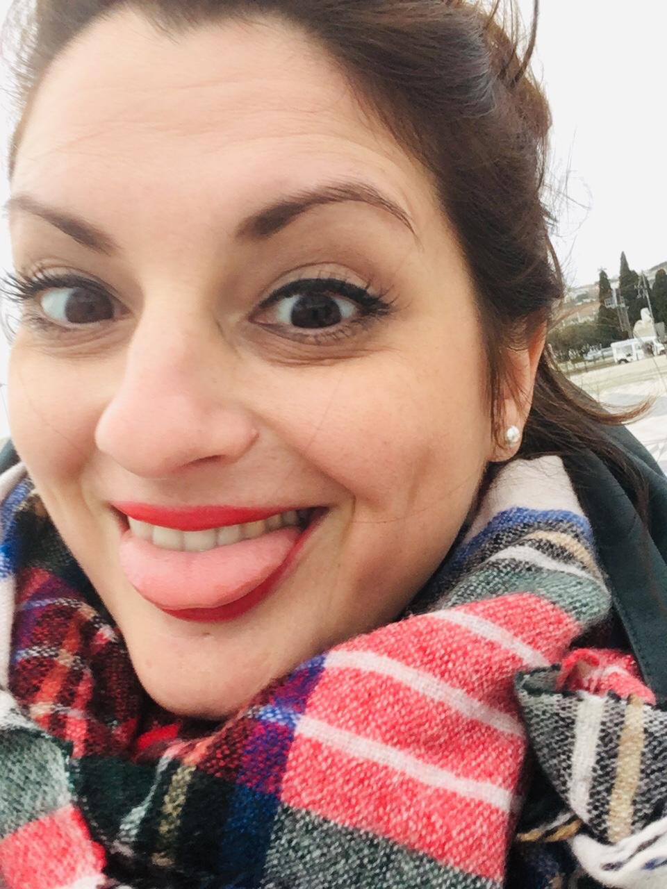 A woman with dark hair and pearl earrings sticking out her tongue, smiling and wearing a colorful plaid scarf. She is outdoors with a cloudy sky and some trees and buildings in the background.