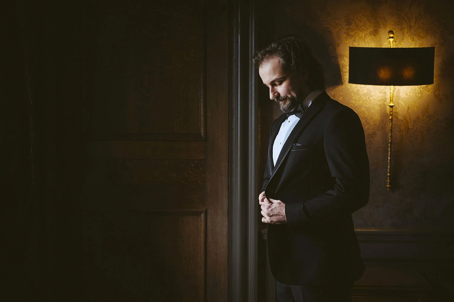 Craythorne Hall wedding: groom in a tuxedo and bow tie looking down thoughtfully beside a warm wall lamp and dark panelled door.