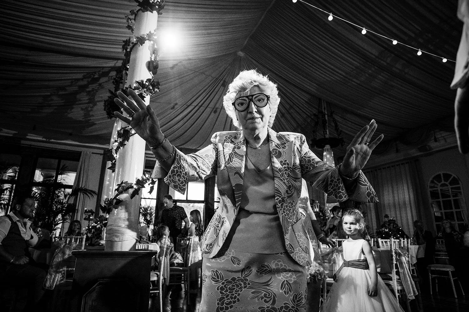 Beamish Woods wedding: elderly guest in glasses dancing with her arms raised as a flower girl looks on, black and white
