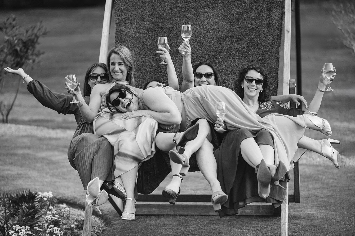 Newton Hall wedding: group of guests in sunglasses laughing as they pile across a bench with glasses raised, in black and white.
