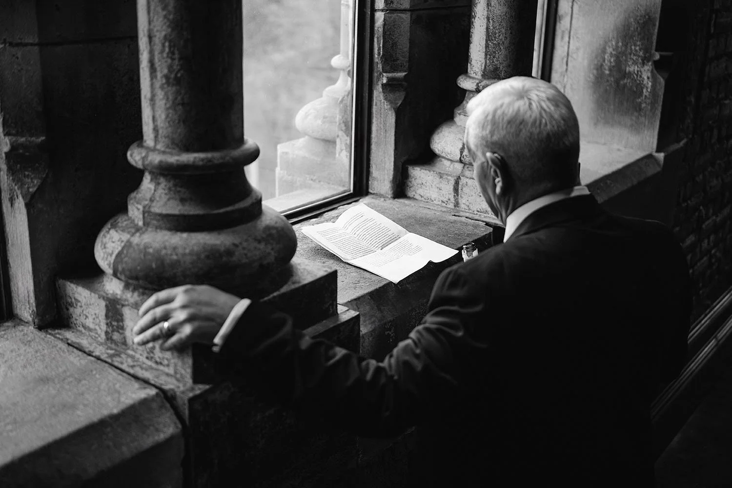 Dalton Pump House wedding: older man in a suit reading a letter by a stone window with a carved pillar, in black and white.