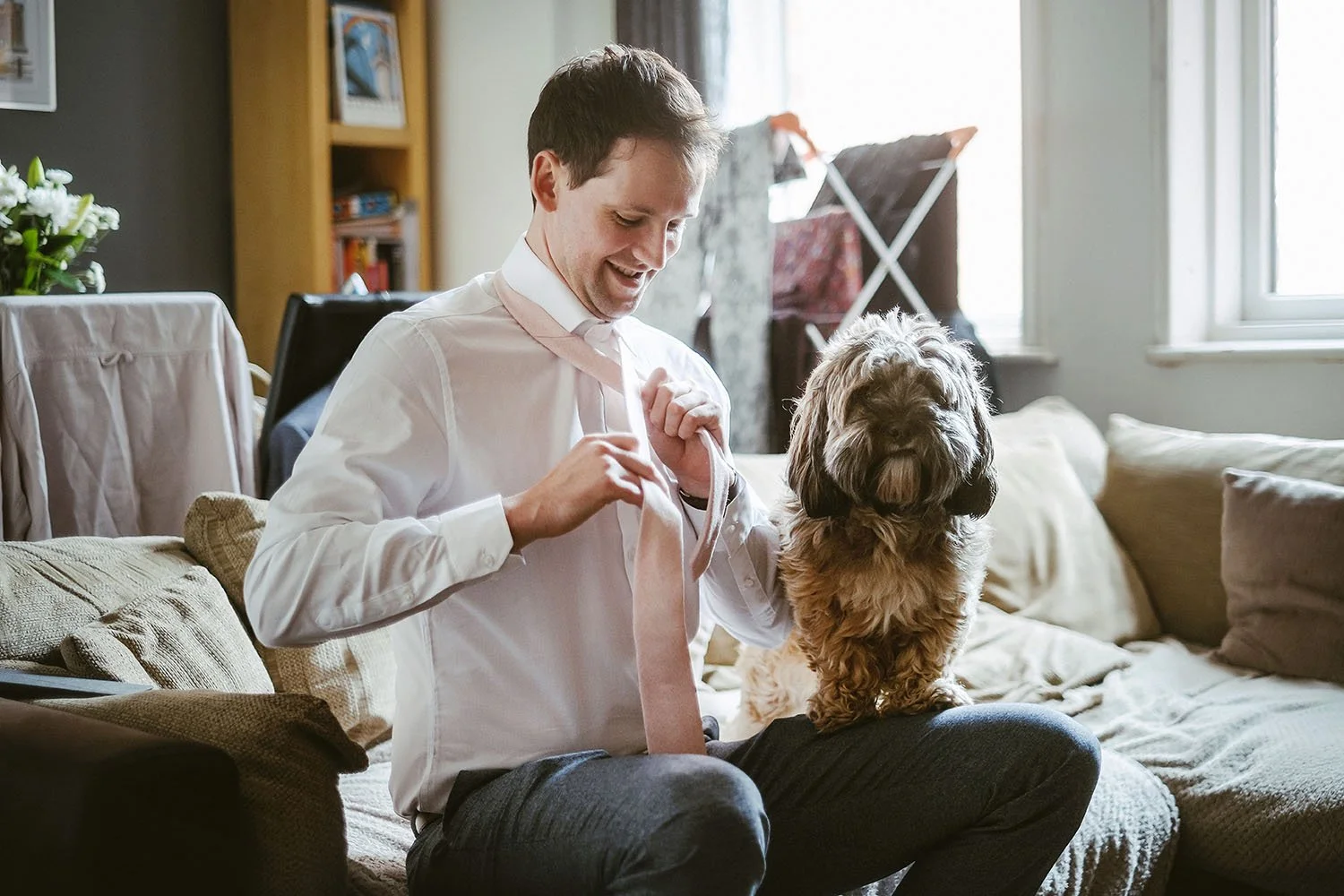 Anarchy Brewery wedding: groom smiling as he ties his pink tie on the sofa beside a shaggy dog.
