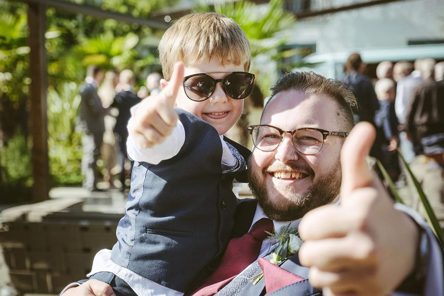 Le Petit Chateau wedding: groom holding a young boy in sunglasses as both give a thumbs-up to the camera