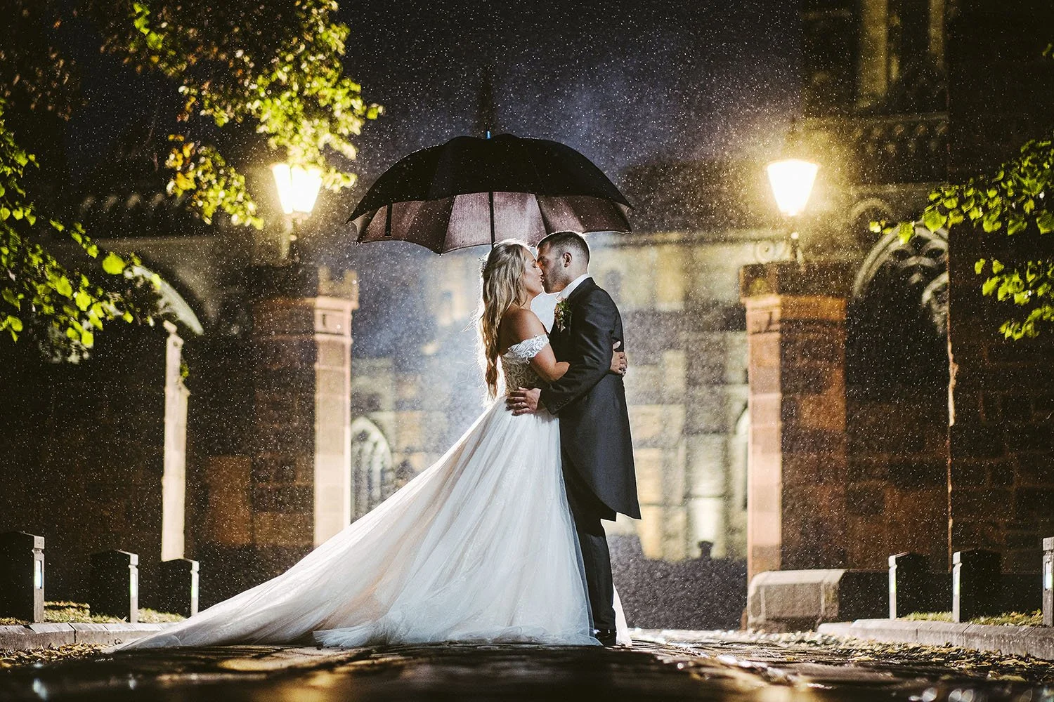 Durham Castle wedding: bride and groom kissing under an umbrella in the rain at night between lit gateposts.