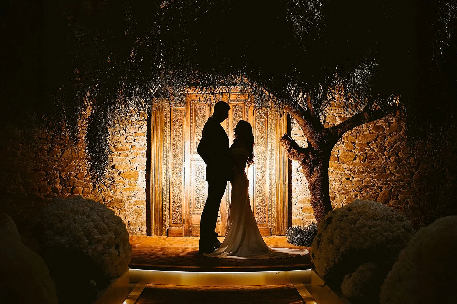 The Croft Hotel wedding: bride and groom in silhouette in front of a lit carved wooden door beneath an olive tree at night.