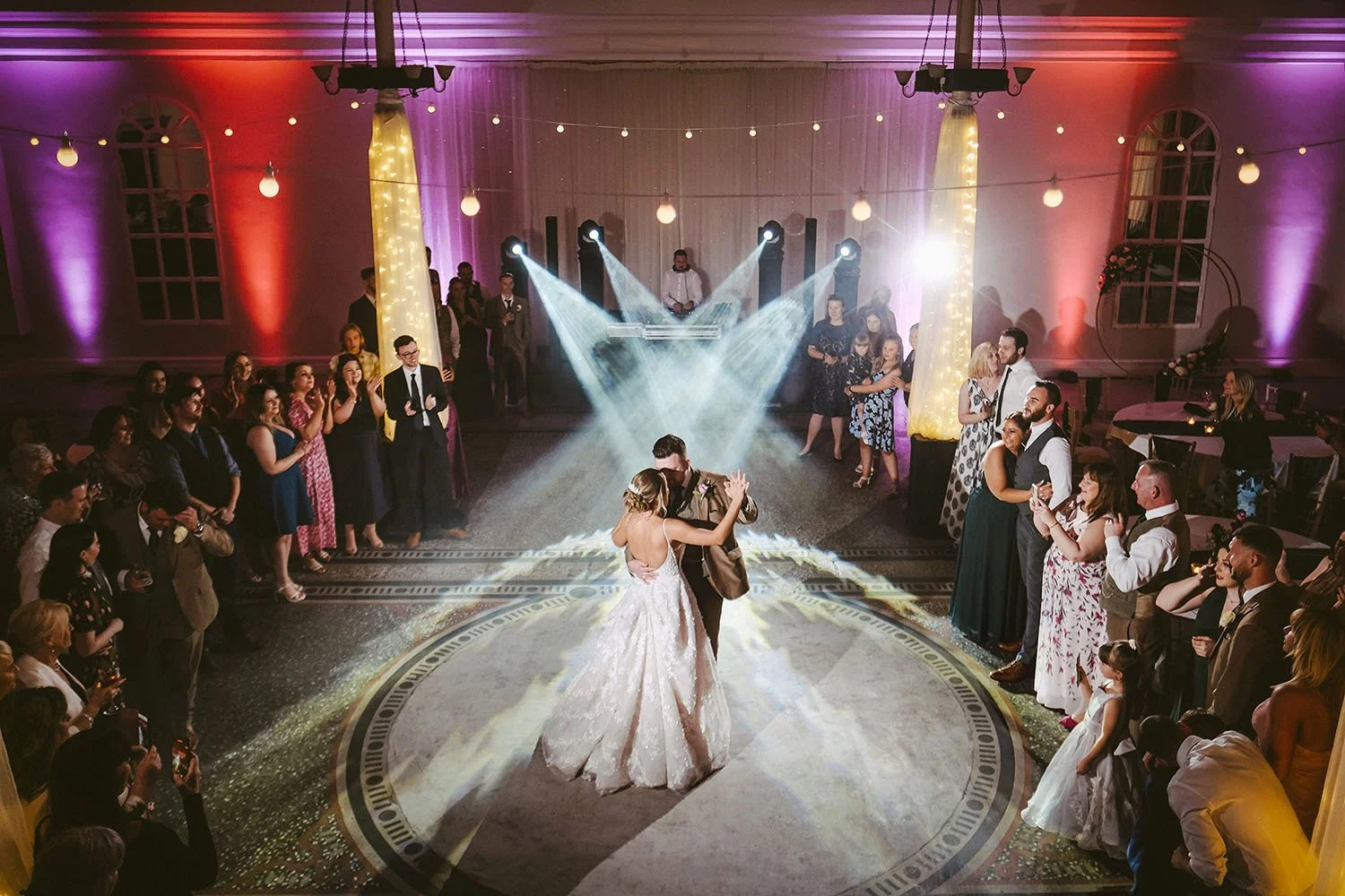 Bride and groom dancing under spotlights in a hall lit in red and purple as guests look on.