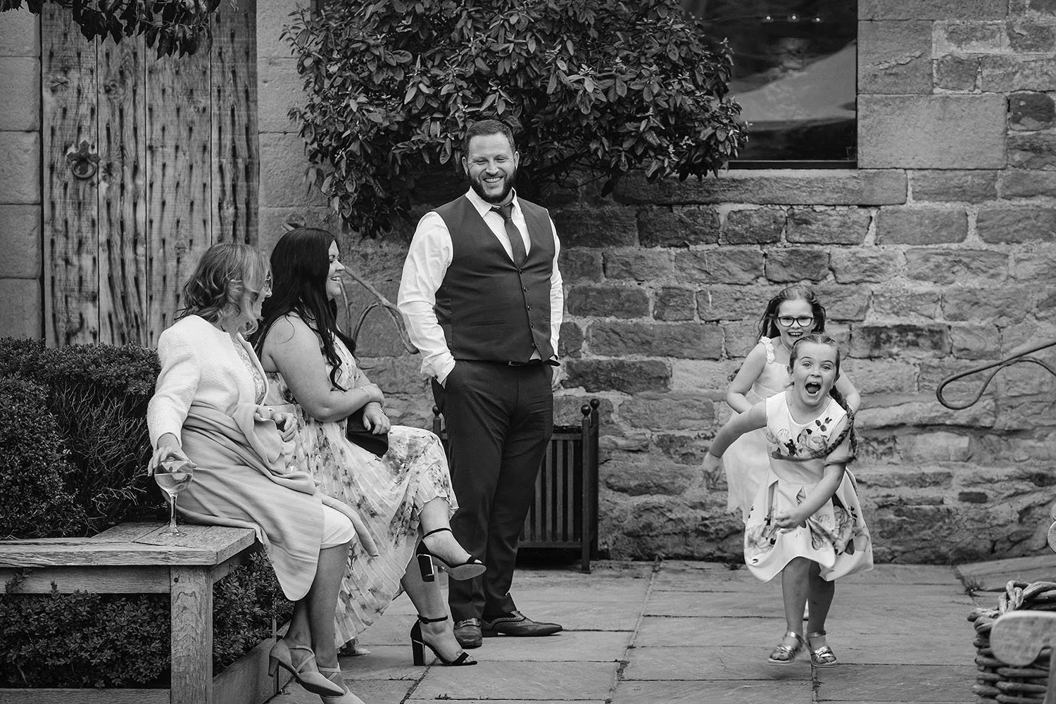 Healey Barn wedding: guests relaxing on a bench as two young girls run through a stone courtyard, in black and white.