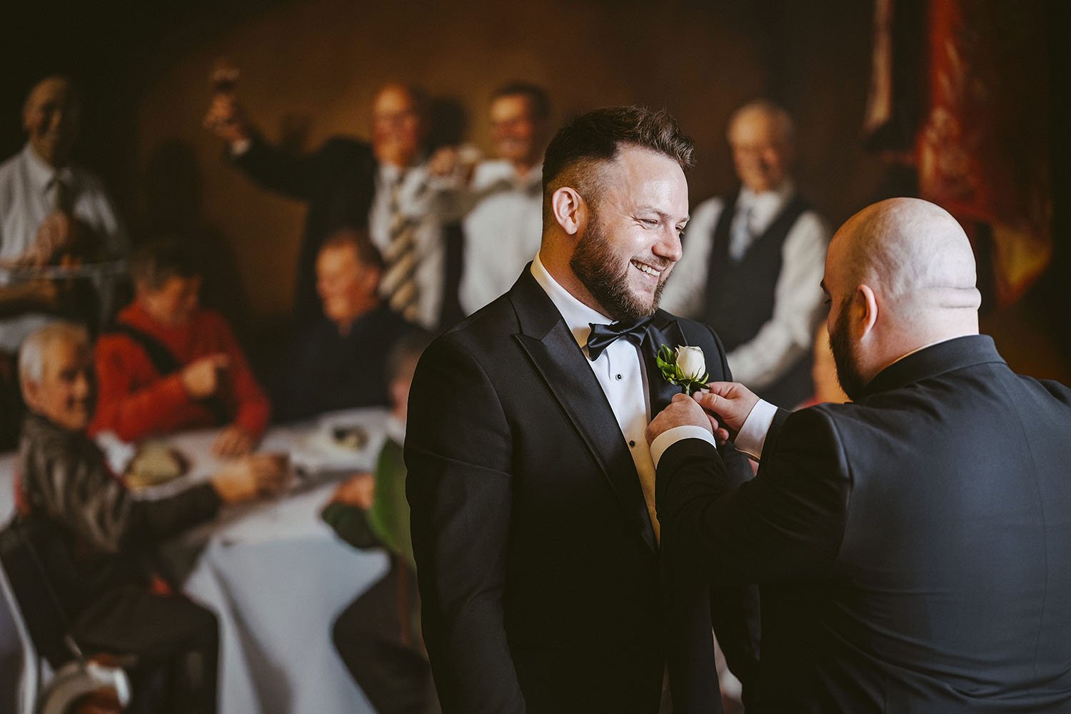 Hardwick Hall wedding: groom smiling in a dark suit as a family member pins his buttonhole, with guests gathered behind.