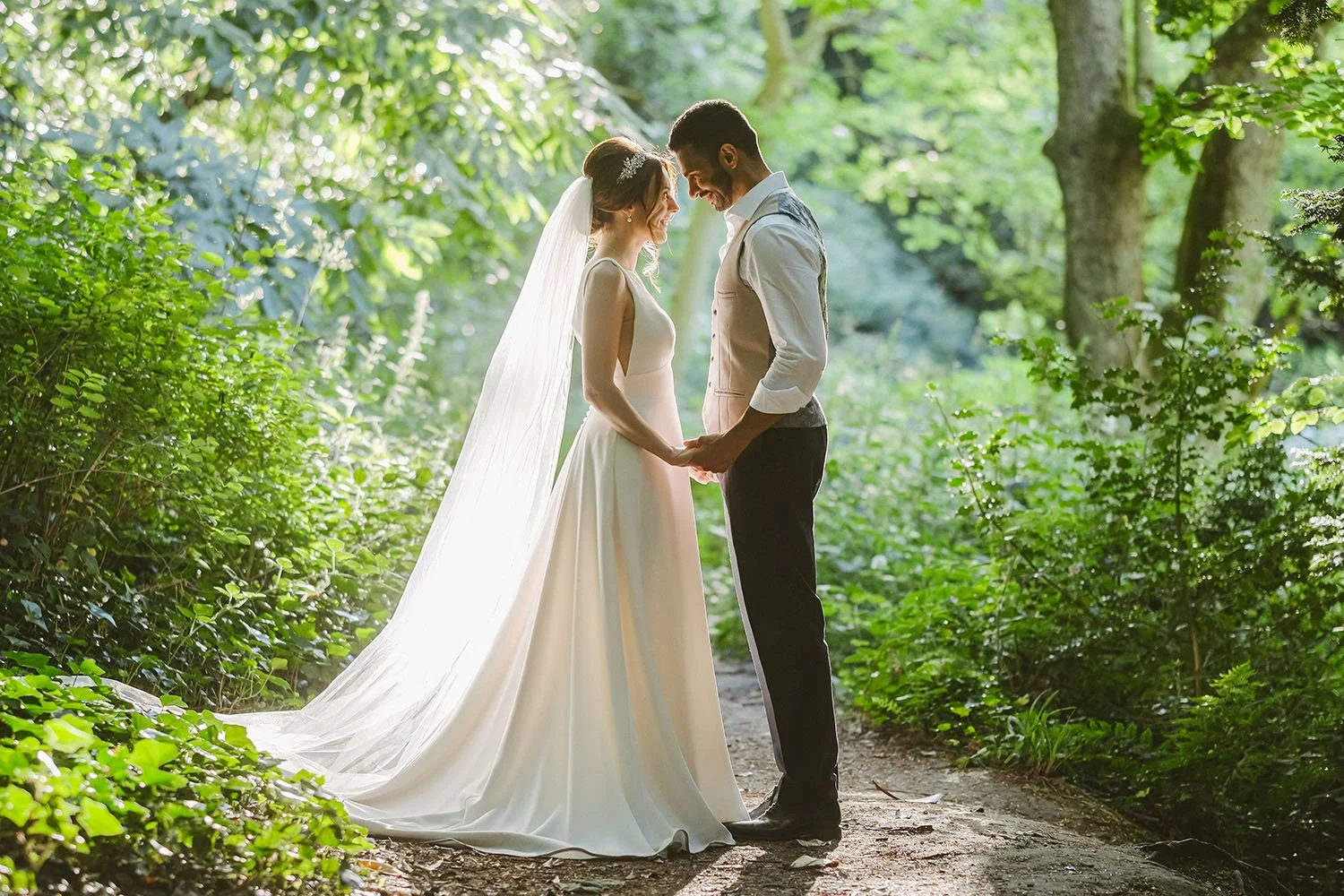 Jesmond Dene House wedding: bride and groom touching foreheads and holding hands in lush green woodland, her long veil trailing.